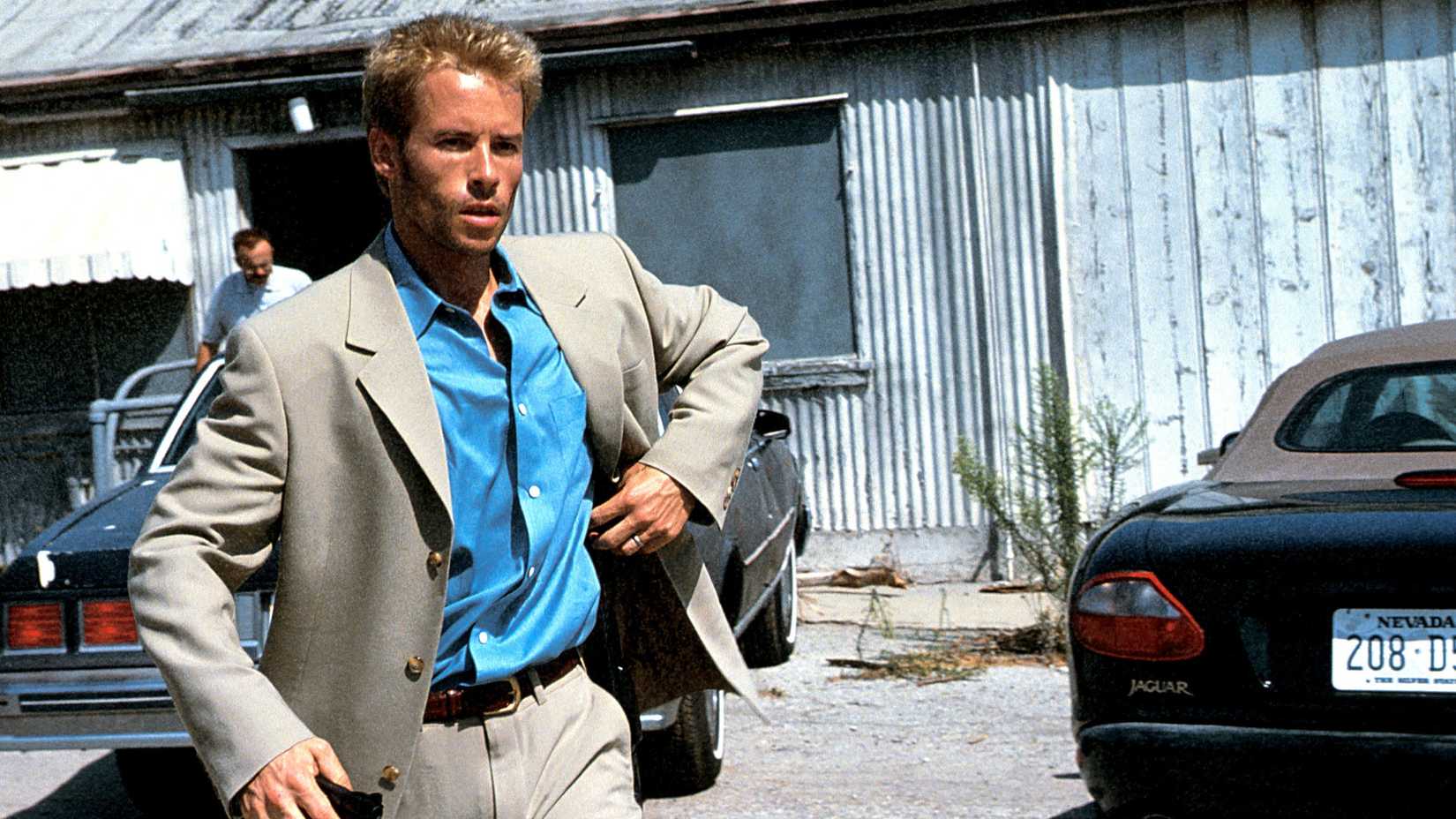 Leonard Shelby (Guy Peace) a blonde man wearing a suit and blue shirt stands outside a worn down building with two cars parked outside of it in Memento