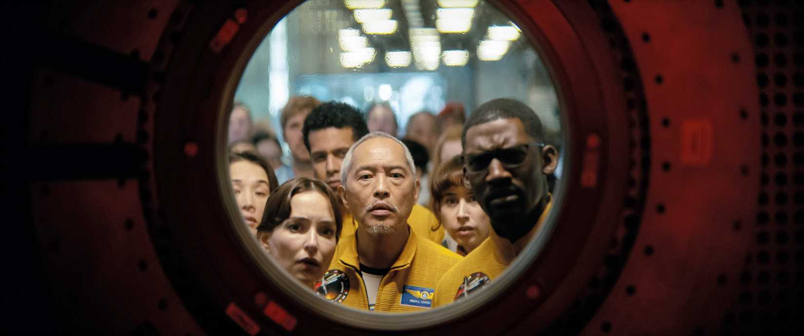 A crowd of people, some in bright yellow jumpsuits, peer through a round porthole into a dark space in Project Hail Mary