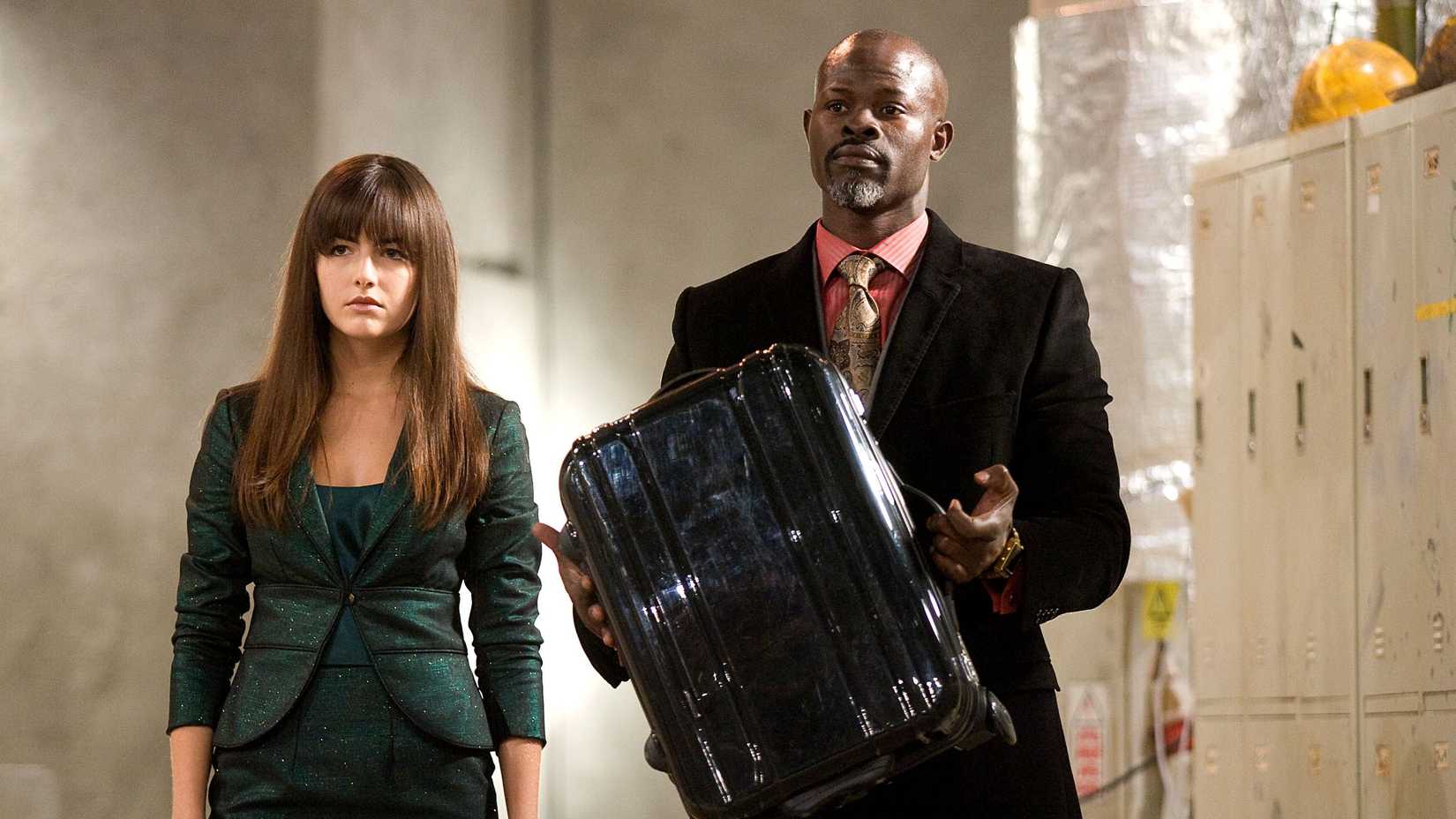 Kira (Camilla Belle) a woman wearing a green suit and Henry Carver (Djimon Hounsou) wearing a black suit and a pink shirt and holding a black suitcase stand near a set of lockers at a construction site in Push