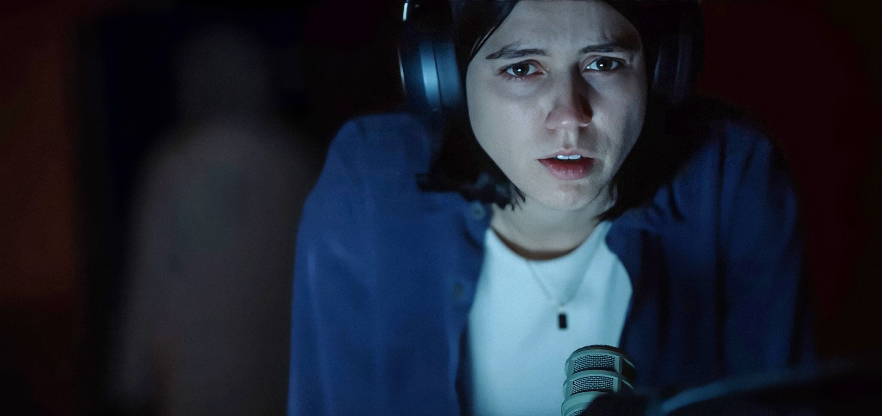 Evy (Nina Kiri), a dark-haired young woman in a white shirt and blue cardigan, wears headphones and looks fearful as she sits in the dark, lit by her laptop's screen, in Undertone