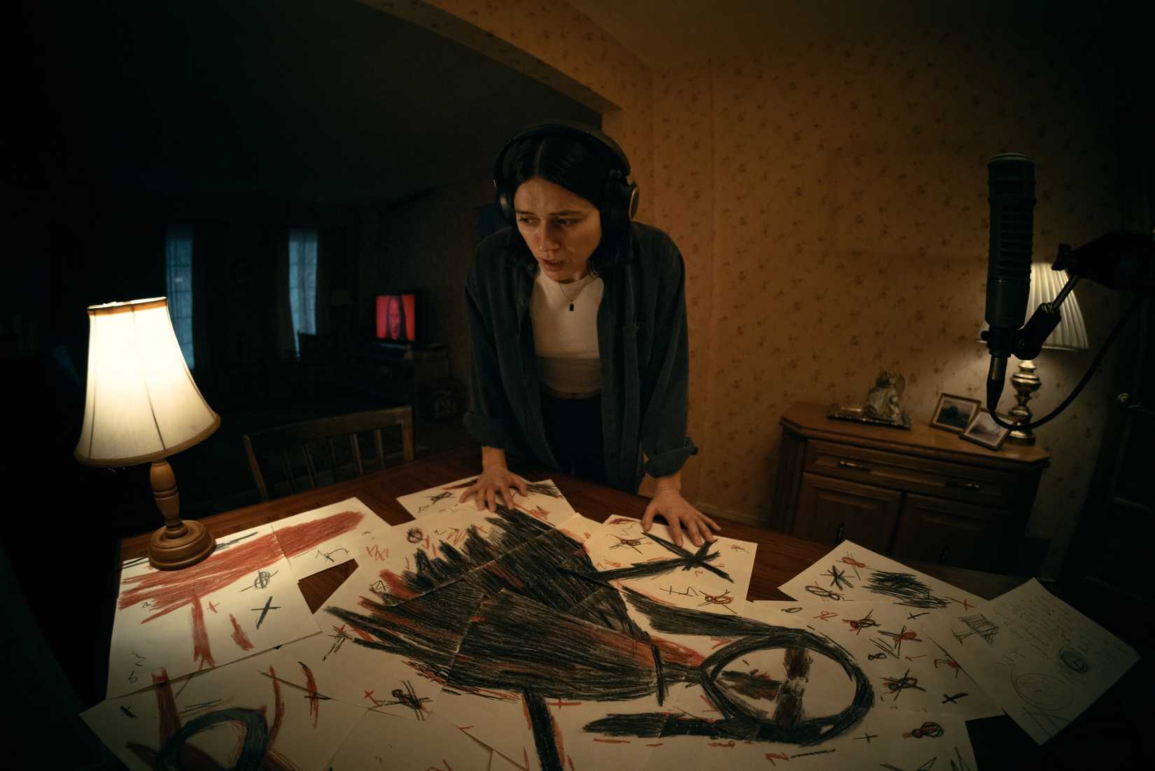 Evy (Nina Kiri), a young woman with dark hair and can headphones, stands in a dark room, leaning over a table covered in sheets of scribbled-on paper assembled in a mosaic to form a black stick-figure-like drawing of a creepy female form. From Undertone.