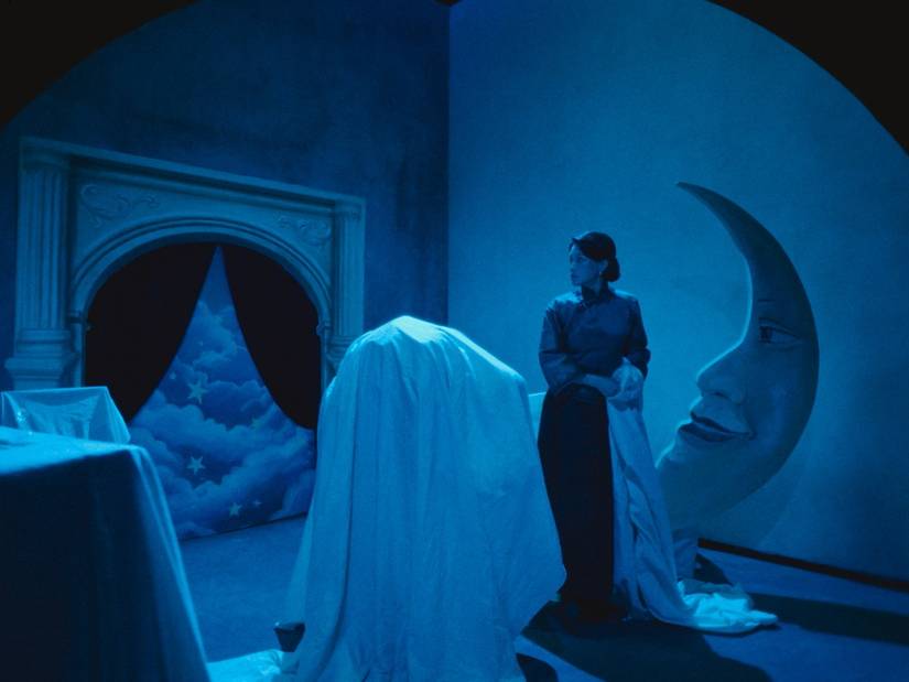 Shu Wi stands amid silent movie props, including a cut-out moon face, some draped in sheets, in Resurrection