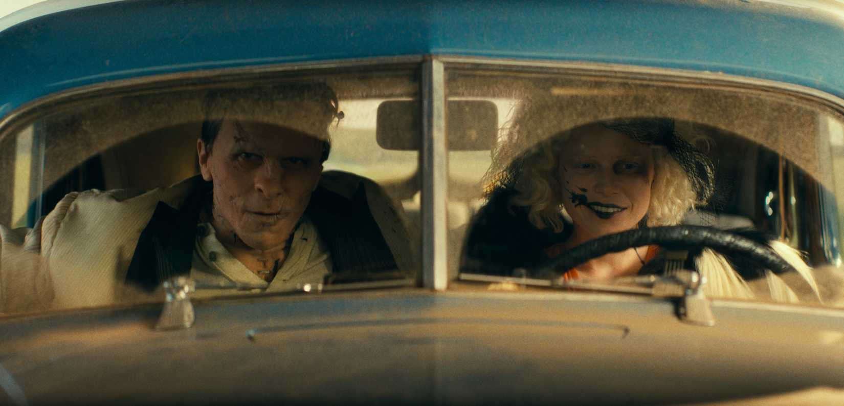 Frank (Christian Bale), aka Frankenstein's monster, a sewn-together man covered in rusty surgical staples, sits in a dusty car alongside Ida (Jessie Buckley), a pale reanimated corpse smeared with black fluid in The Bride!