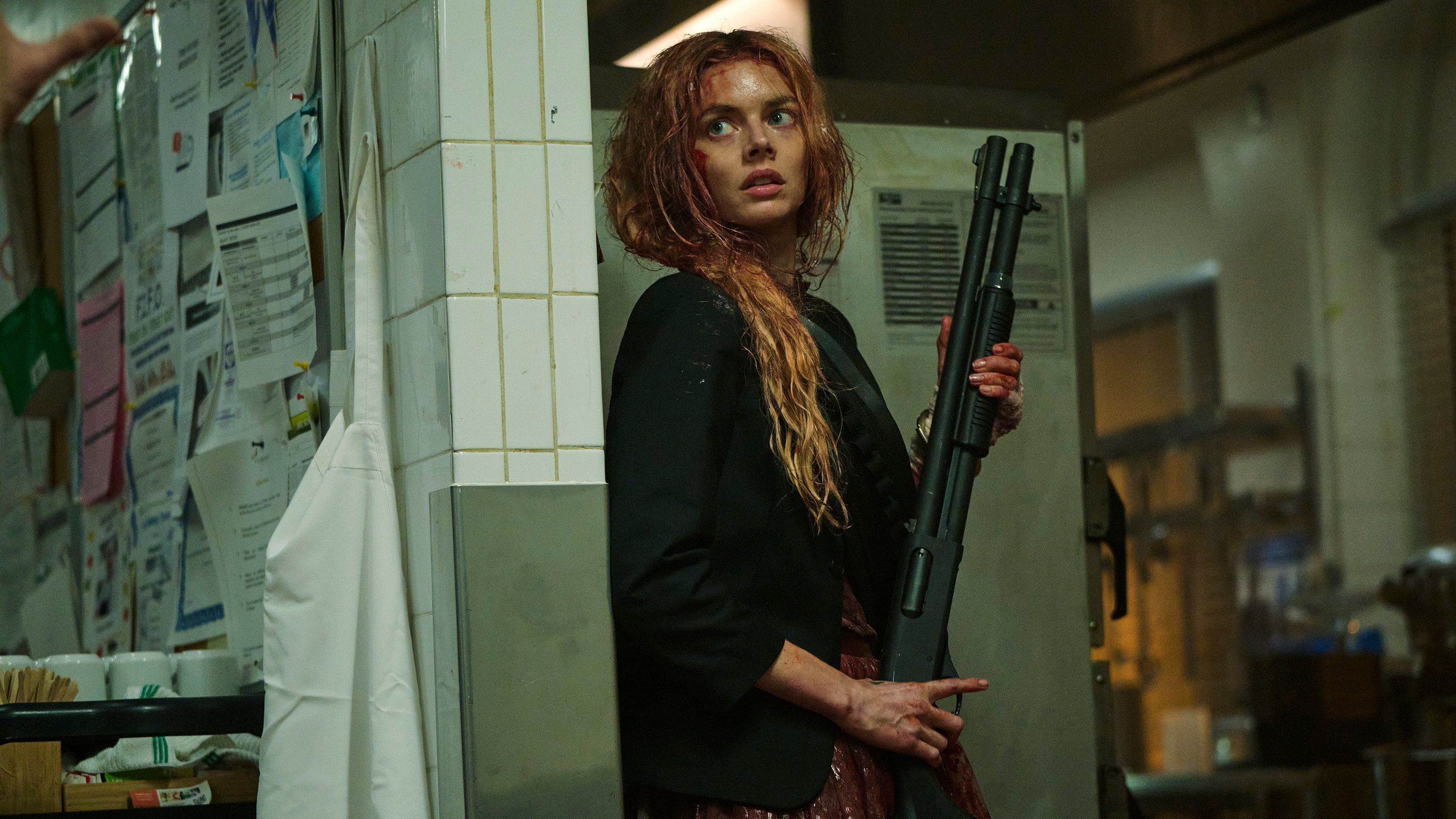 Grace (Samara Weaving), a woman wearing a bloodstained wedding dress and a suit jacket, holds a shotgun while hiding in a hotel kitchen in Ready or Not 2