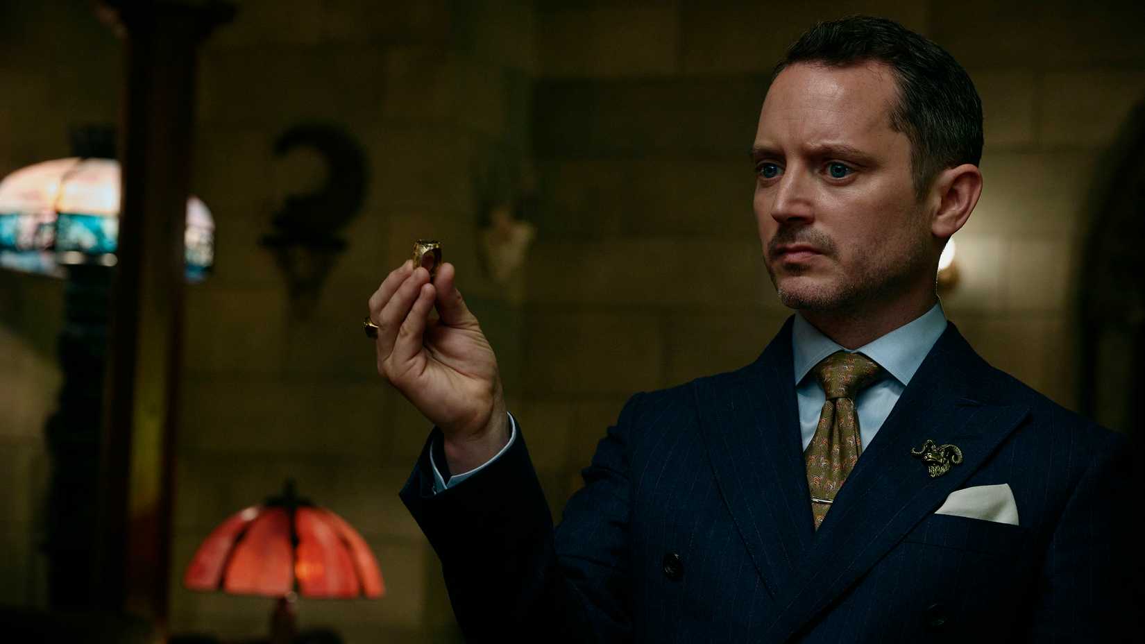The Lawyer (Elijah Wood), a man wearing a blue suit with a goat head lapel pin, holds out a gold ring while standing in a lamplit room in Ready or Not 2.