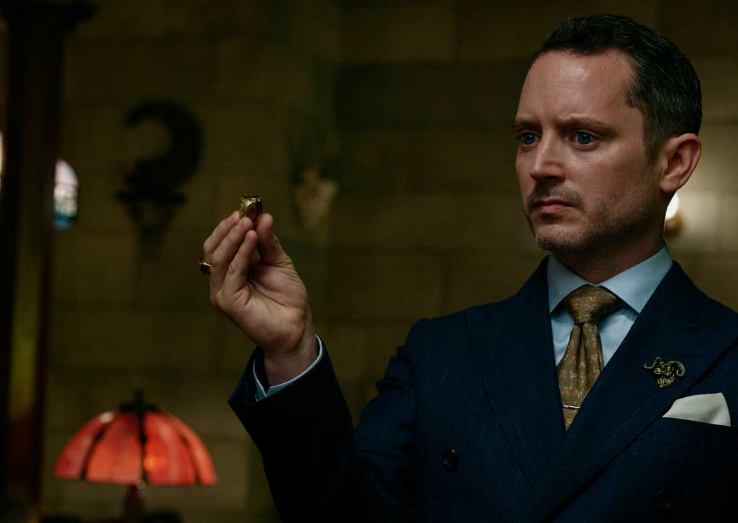 The Lawyer (Elijah Wood), a man wearing a blue suit with a goat head lapel pin, holds out a gold ring while standing in a lamplit room in Ready or Not 2.