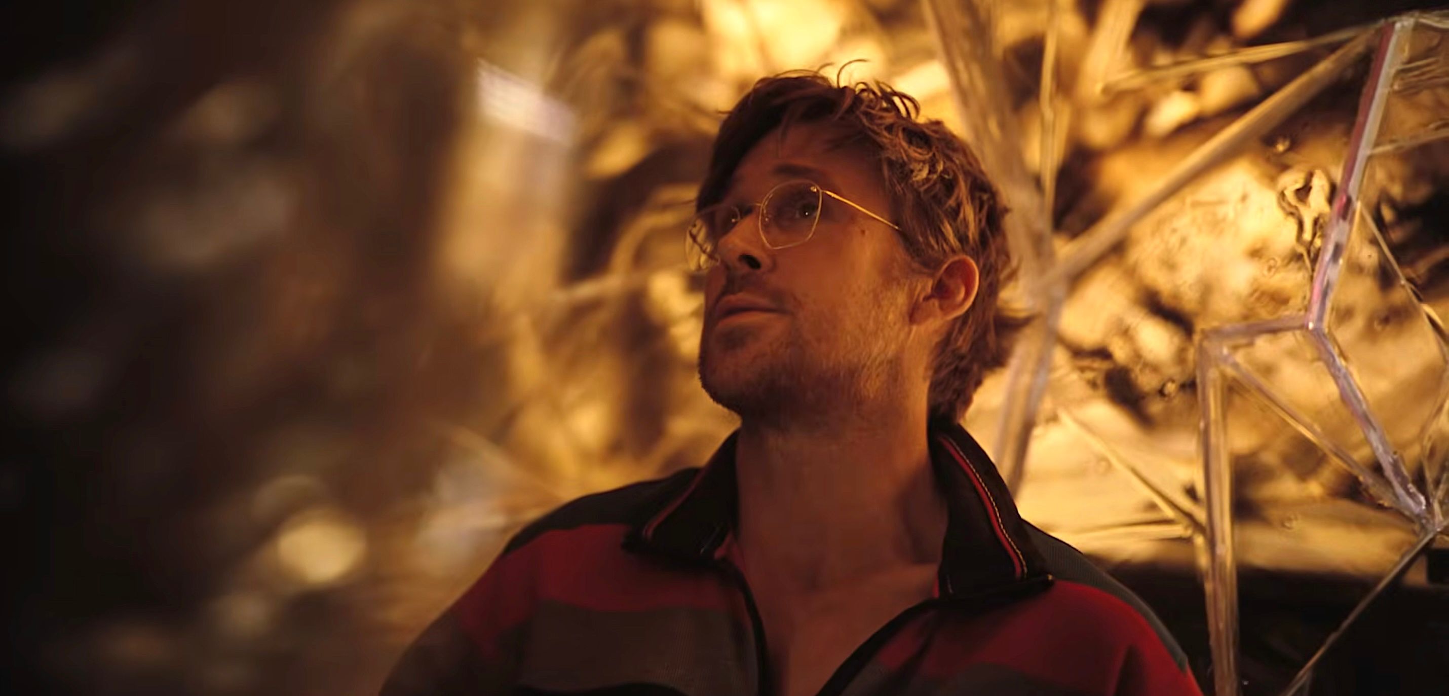 Astronaut Ryland Grace (Ryan Gosling), shaggy-haired, wearing glasses and an orange jumpsuit, in front of a crystal-faceted background
