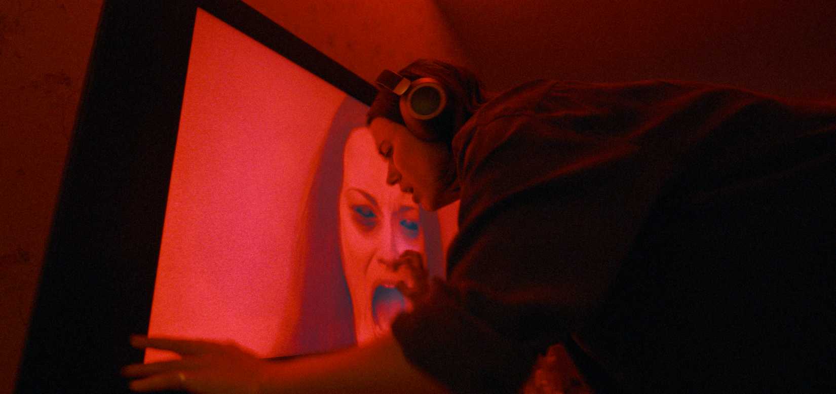 Evy (Nina Kiri), a young woman with dark hair and can headphones, leans over a flatscreen TV displaying a red-lit image of a screaming woman with blacked-out eyes in Undertone