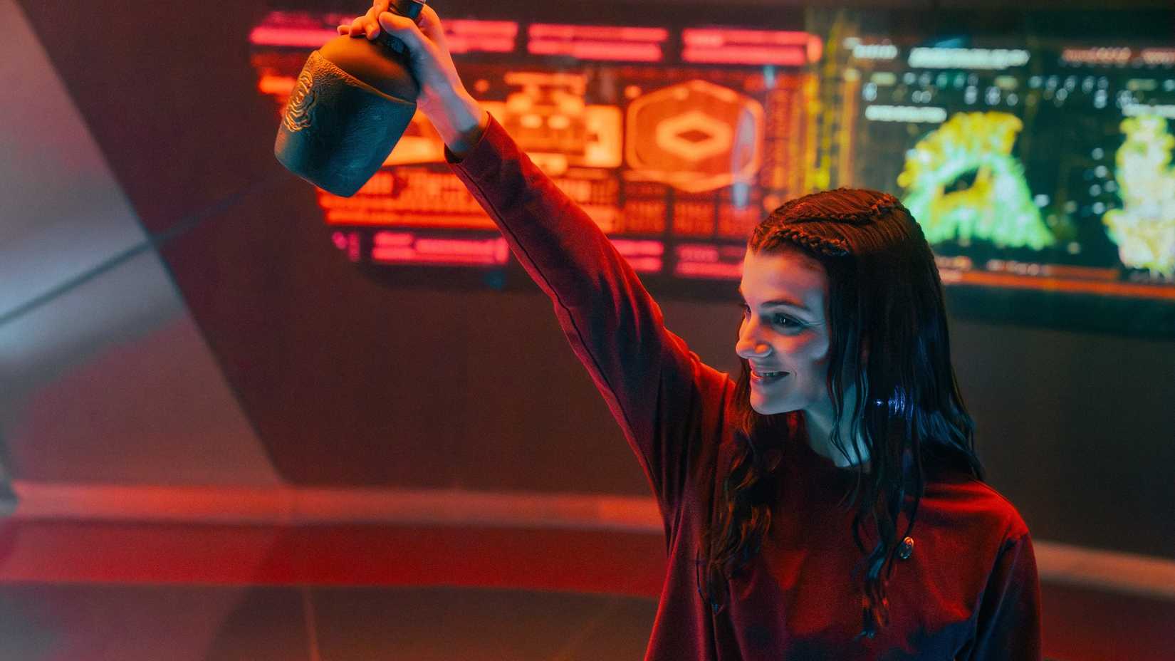 Tarima Sadal (Zoë Steiner) holds up a jug of a strongly alcoholic beverage in front of a panel of screens on the USS Athena