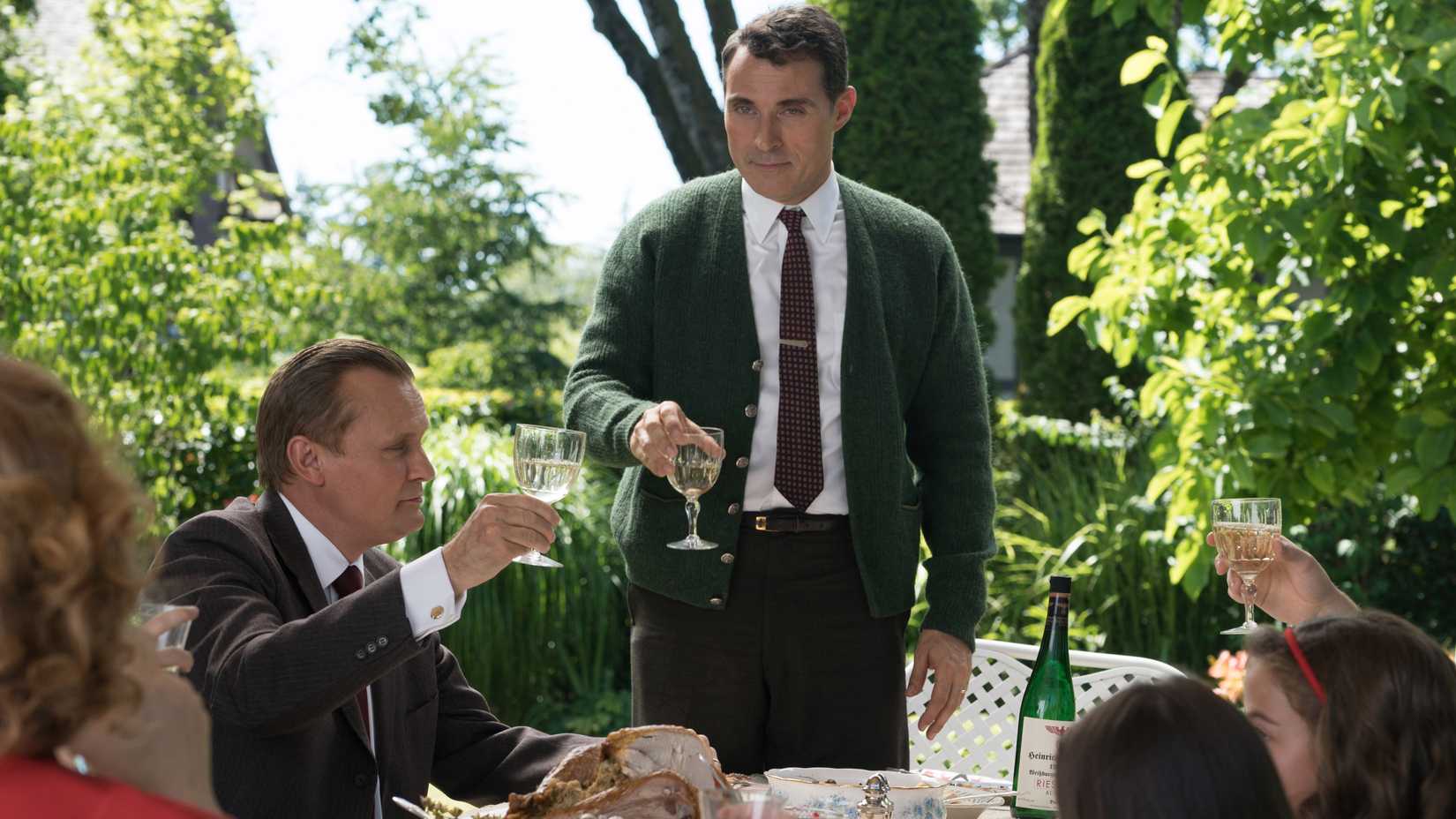 John Smith (Rufus Sewell) holds a glass of wine for a toast as a garden party in The Man in the High Castle