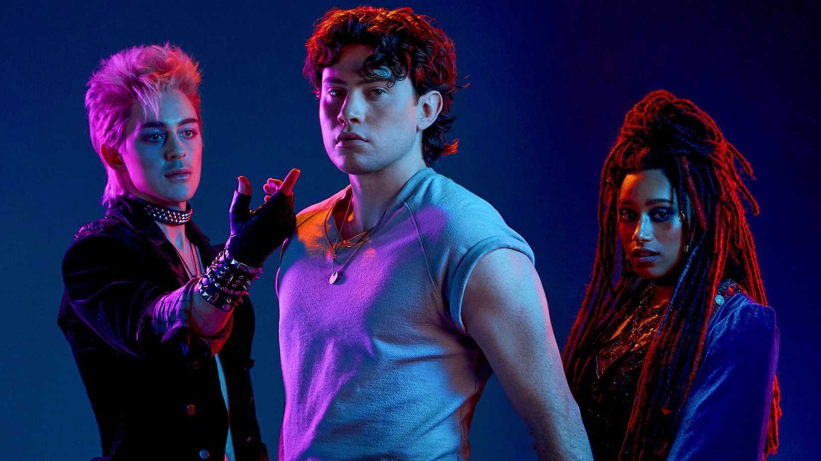 David (Ali Louis Bourzgui) a guy wearing spiky jewelry and a trenchcoat, reaches out to Michael (LJ Benet) a young man in jeans and a gray shirt, while Star (Maria Wirries), a woman wearing a velvety robe, stands behind him in The Lost Boys musical