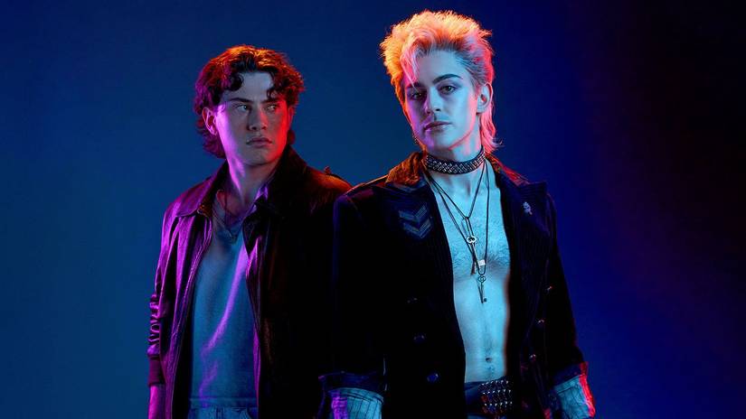 Michael (LJ Benet) a young man wearing a leather jacket, looks at David (Ali Louis Bourzgui) a shirtless man wearing a trenchcoat and combat boots in The Lost Boys musical