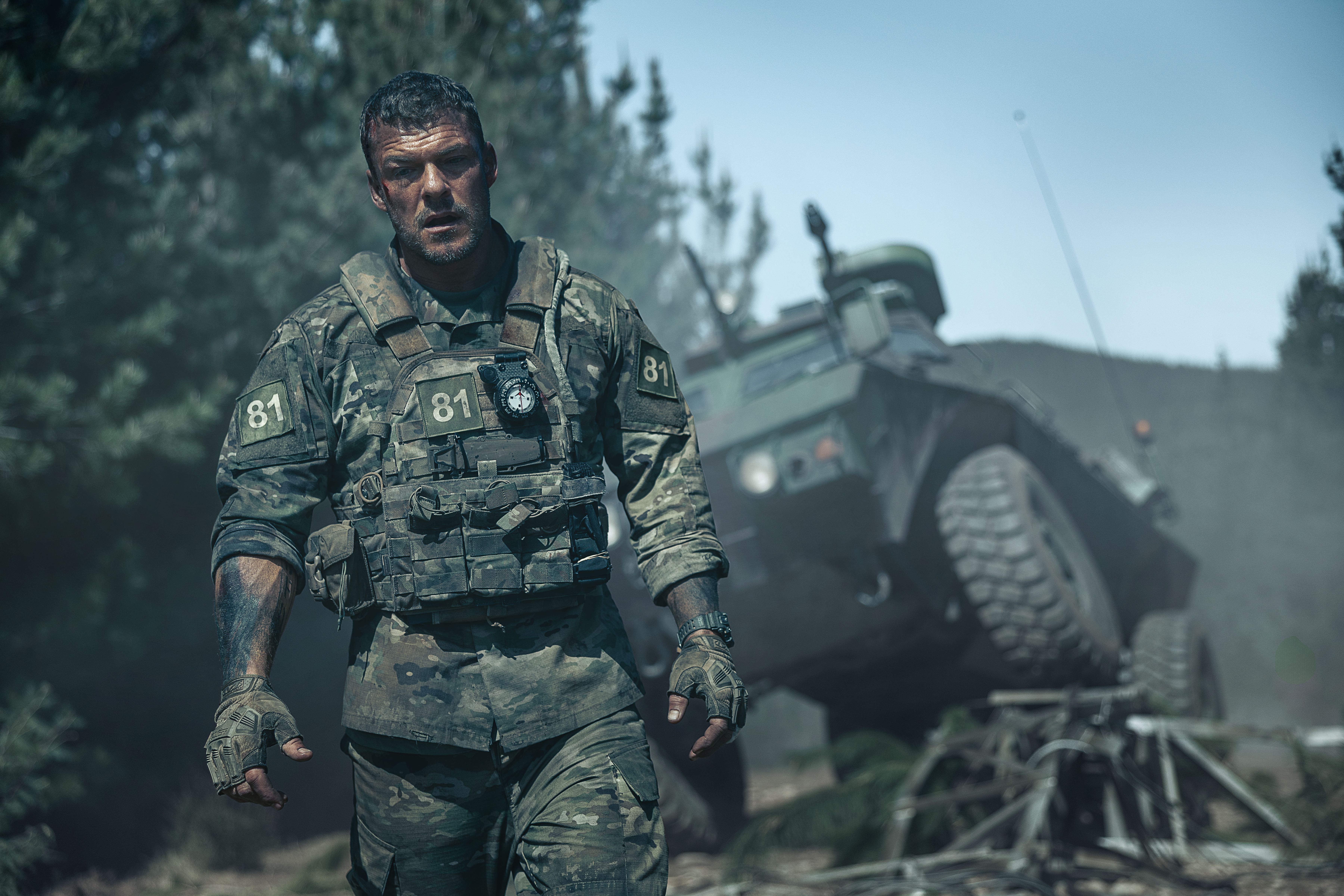 81 (Alan Ritchson), an Army sergeant in grimy fatigues, covered with mud and blood, stands in front of a wrecked army vehicle in War Machine