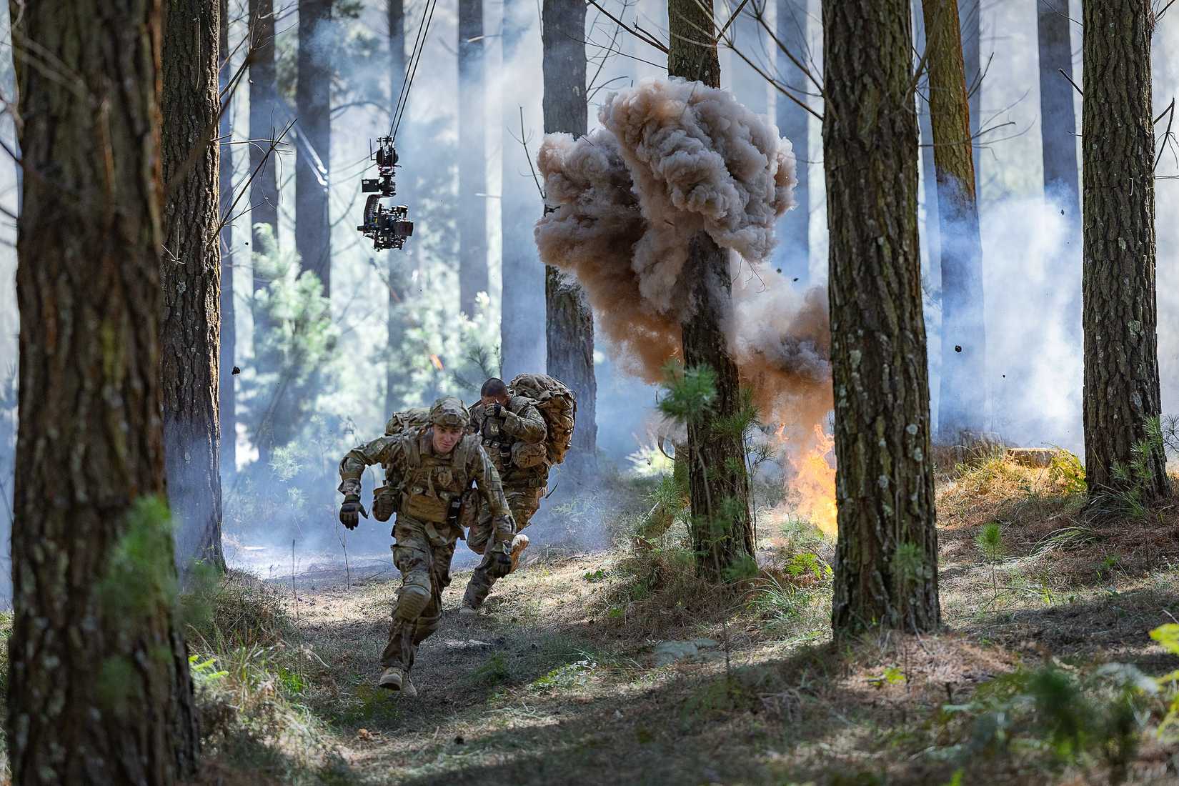 Actors dressed as Army trainees run through trees as explosions are set off and a camera hovers on wires overhead on the set of War Machine