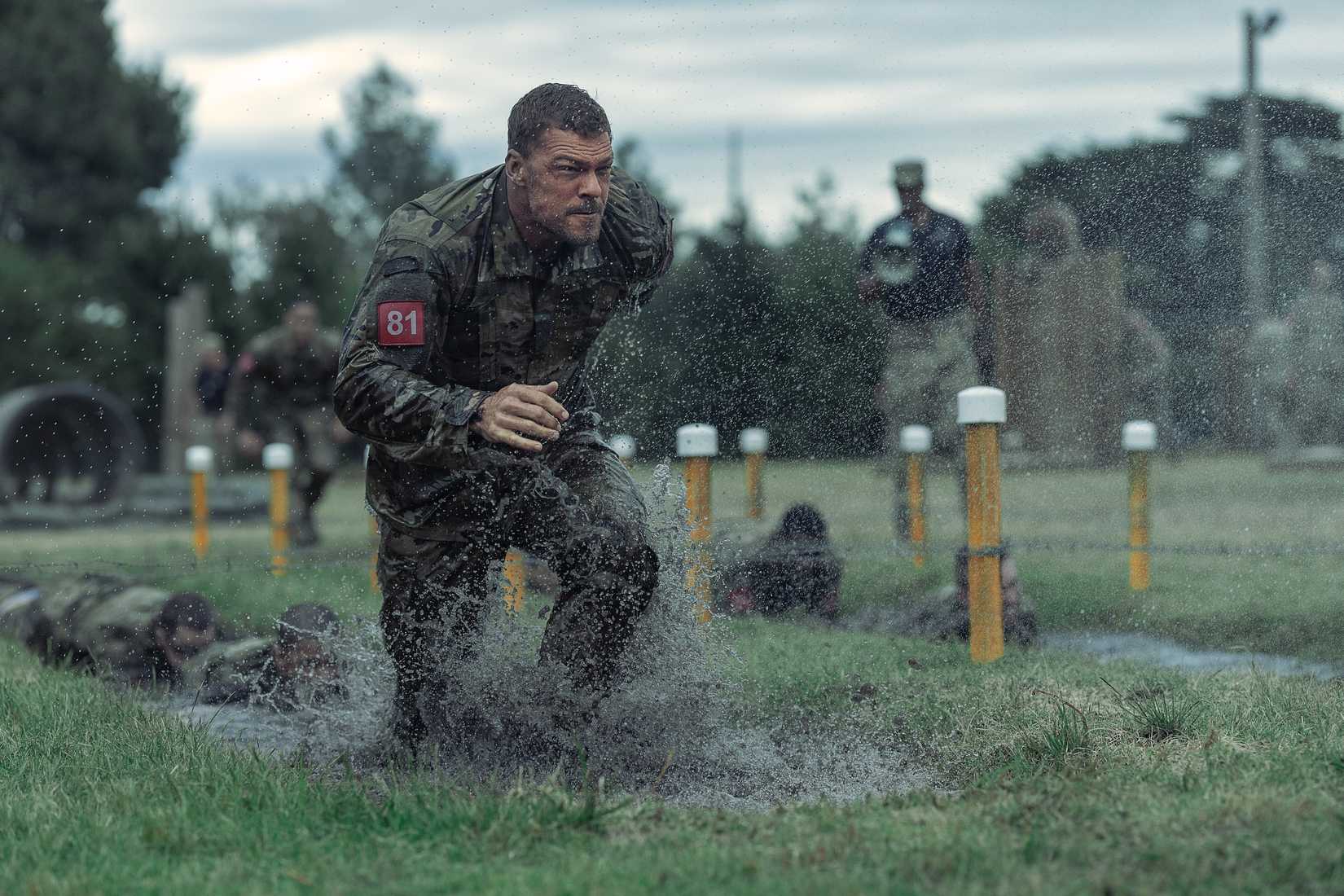 Army sergeant 81 (Alan Ritchson), soaking wet in fatigues and red identifying number, runs across a sodden field amid yellow lane marker posts in War Machine