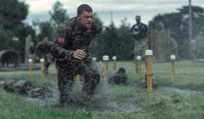 Army sergeant 81 (Alan Ritchson), soaking wet in fatigues and red identifying number, runs across a sodden field amid yellow lane marker posts in War Machine