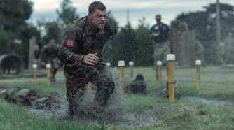Army sergeant 81 (Alan Ritchson), soaking wet in fatigues and red identifying number, runs across a sodden field amid yellow lane marker posts in War Machine