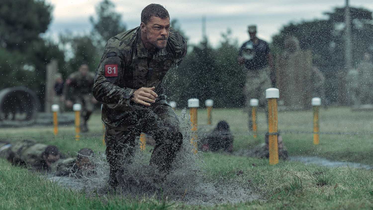 Army sergeant 81 (Alan Ritchson), soaking wet in fatigues and red identifying number, runs across a sodden field amid yellow lane marker posts in War Machine