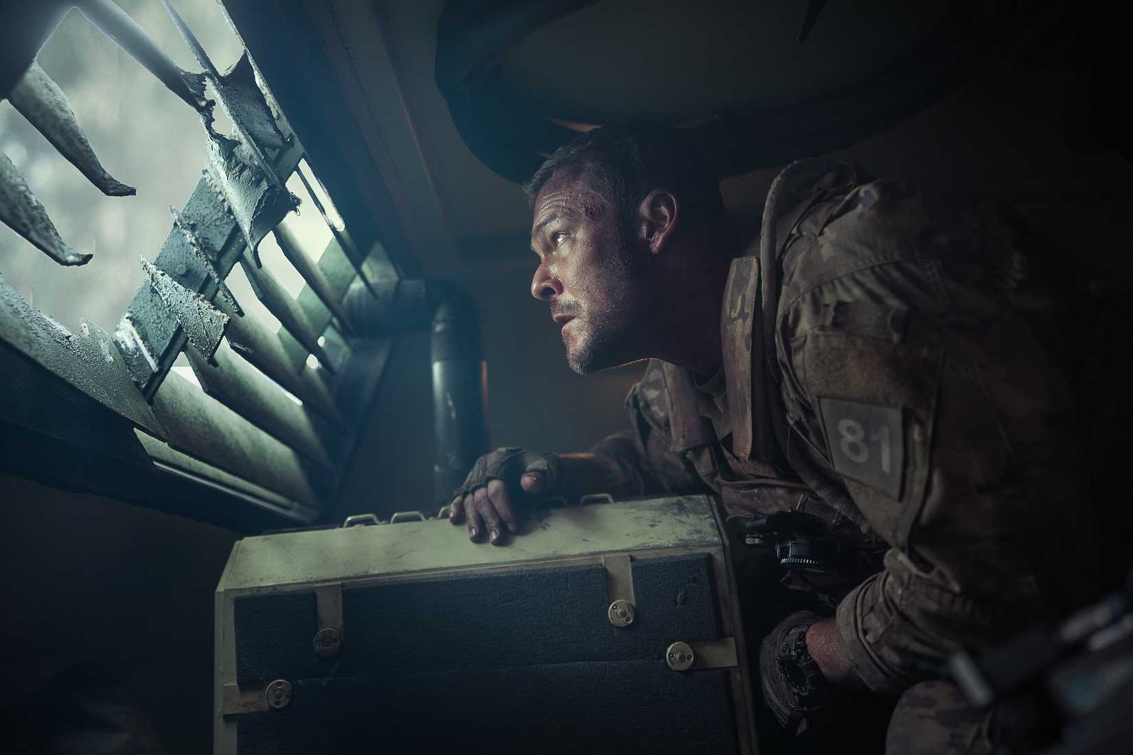Army sergeant 81 (Alan Ritchson), looking unnerved, stands hunched over inside an armored truck and peers out through a louvered window in War Machine