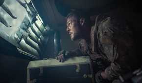 Army sergeant 81 (Alan Ritchson), looking unnerved, stands hunched over inside an armored truck and peers out through a louvered window in War Machine
