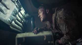 Army sergeant 81 (Alan Ritchson), looking unnerved, stands hunched over inside an armored truck and peers out through a louvered window in War Machine