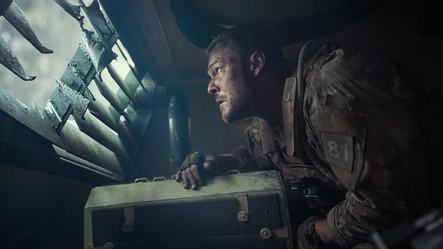 Army sergeant 81 (Alan Ritchson), looking unnerved, stands hunched over inside an armored truck and peers out through a louvered window in War Machine