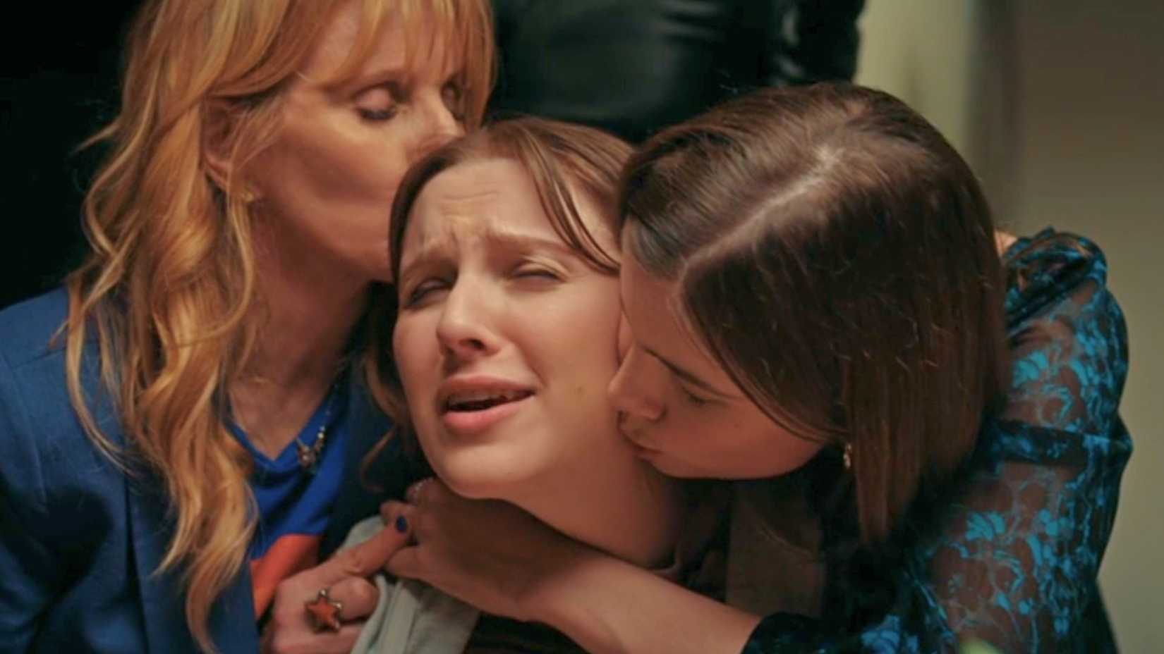 An image from D(e)ad. It shows a blonde older woman kissing the head of her daughter, who is also being hugged by her sister. The daughter in the middle looks annoyed.