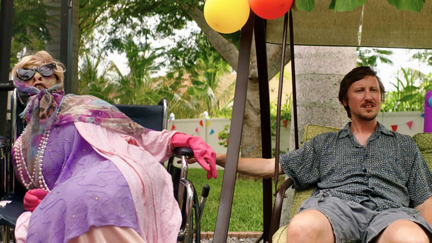 An image featuring Johnny Pemberton in the action-comedy film, Mermaid. It shows Doug (Johnny) sitting down, his hand on the arm of a wheelchair. A mermaid, dressed in old-timey clothing with sunglasses and a bad wig, sits next to him.