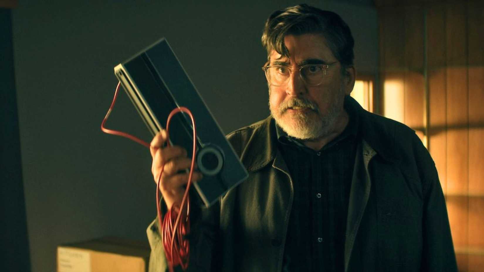 Alfred Molina as Sam Cooper in The Boroughs. He holds up a radio-like device, with red wires.