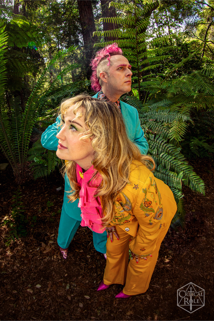 Taliesin Jaffe and Ashley Johnson stand side by side. Ashley, wearing a bedazzled yellow suit, looks to the left. Taliesin, in a blue suit, stares off to the right.