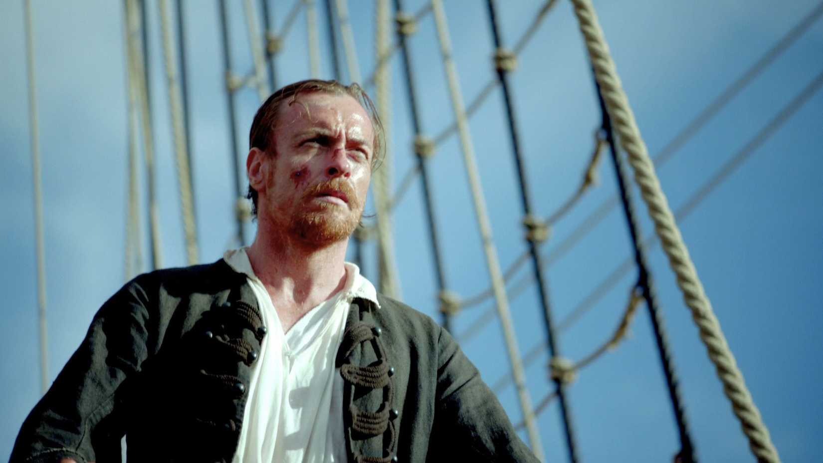 James Flint (Toby Stephens) is bloodied and bruised. His hair is tied up in a short ponytail and he has a majestic beard and goatee. He looks rugged and weary.