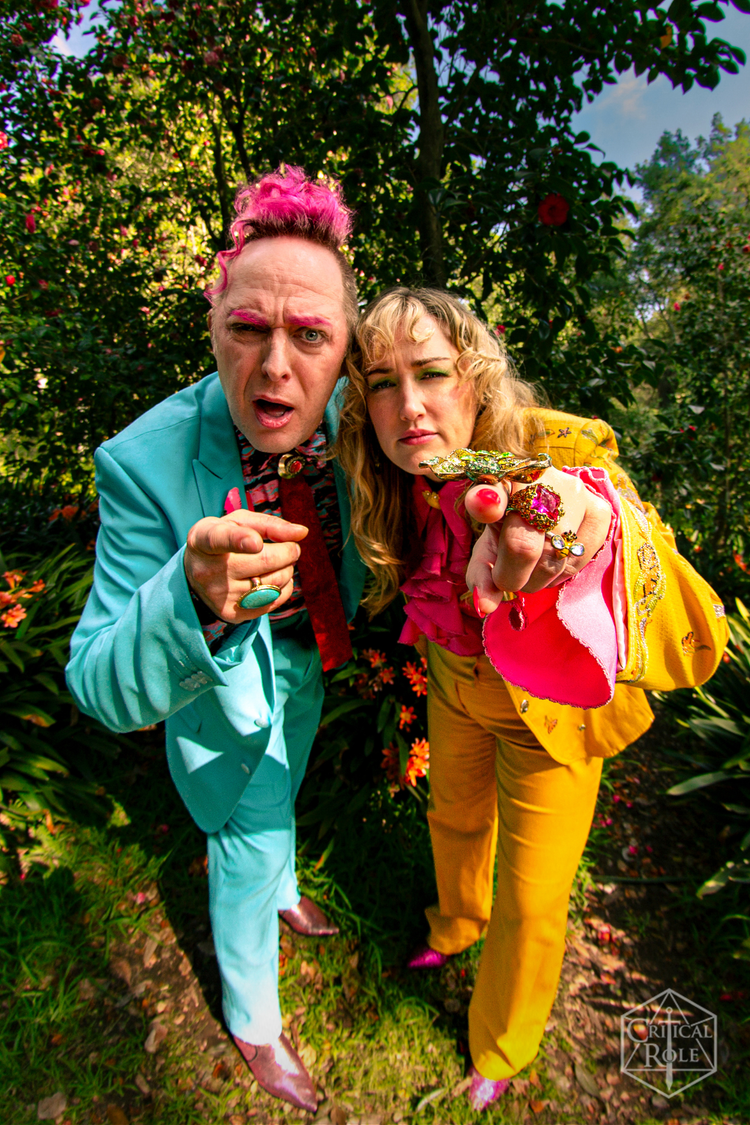 An image featuring Taliesin Jaffe and Ashley Johnson. Ashley wears a yellow suit, and Taliesin a blue. They point at the camera.