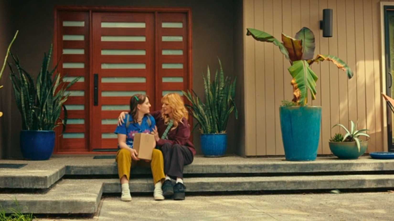 An image from D(e)ad. It shows Tillie, a young woman, in a blue shirt and yellow pants sitting next to a blonde woman in red and black. They are holding one another.