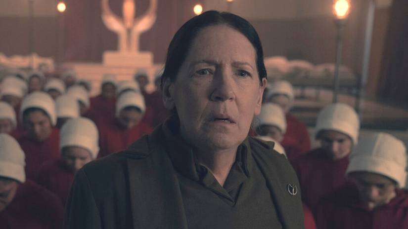 Aunt Lydia (Ann Dowd), an older woman in dark green, looks drained and worried in The Handmaid's Tale. Behind her, a crowd of handmaids — women in dark red robes and white wimples — bow their heads.
