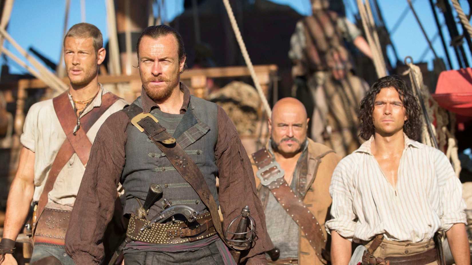 An image from Black Sails. It shows Toby Stephens as Captain James Flint. He is flanked by three men.