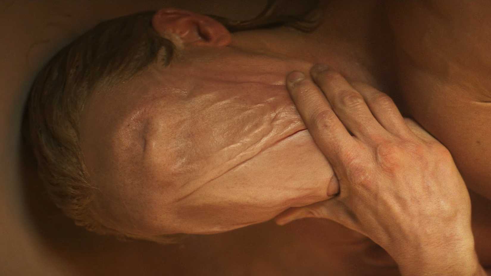 A closeup shot of Clayface, with his hand on his chin and an unbroken flap of skin from forehead to chin, from the movie's first teaser trailer