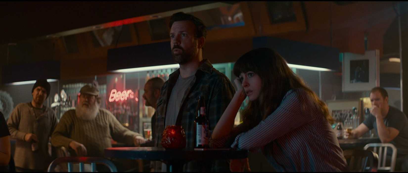 Gloria (Anne Hathaway) and Oscar (Jason Sudeikis) watch an offscreen television from a bar table in a scene from Colossal