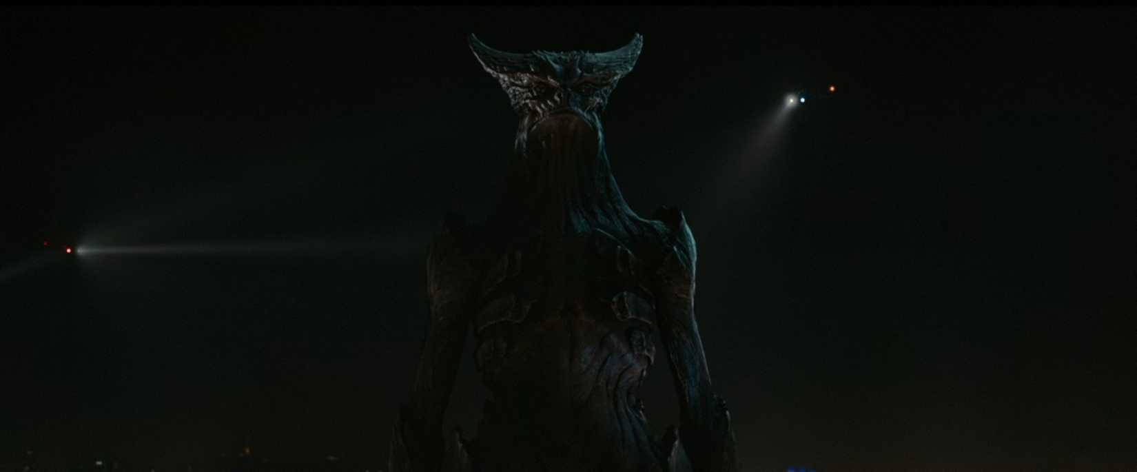 A center-frame shot, in front of a darkened sky, of a towering but humanoid kaiju, the avatar for Anne Hathaway's character in Colossal.