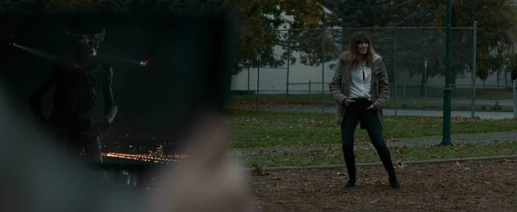 In a scene from Colossal (2017), Gloria (Anne Hathaway) stands in a park, in focus but in the background of the image, posing in unison with a kaiju creature seen in the foreground of the image on a tablet, establishing their bizarre connection.
