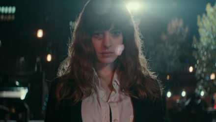 Anne Hathaway, playing Gloria, faces the camera in a center-frame medium close-up with light glaring behind her, lighting up the Seoul night sky, in a scene from the 2017 film Colossal.