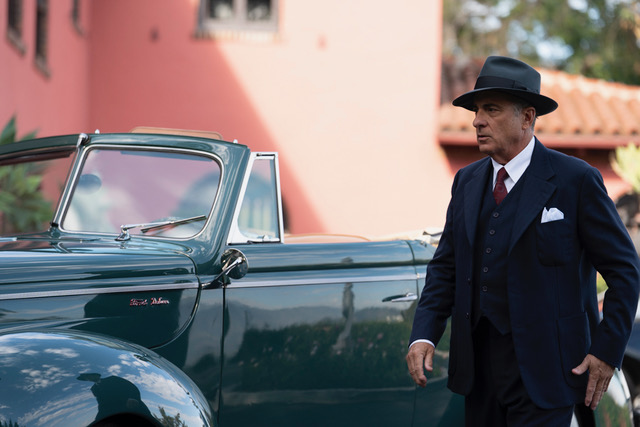 A man in a suit and fedora walking past a shiny classic car
