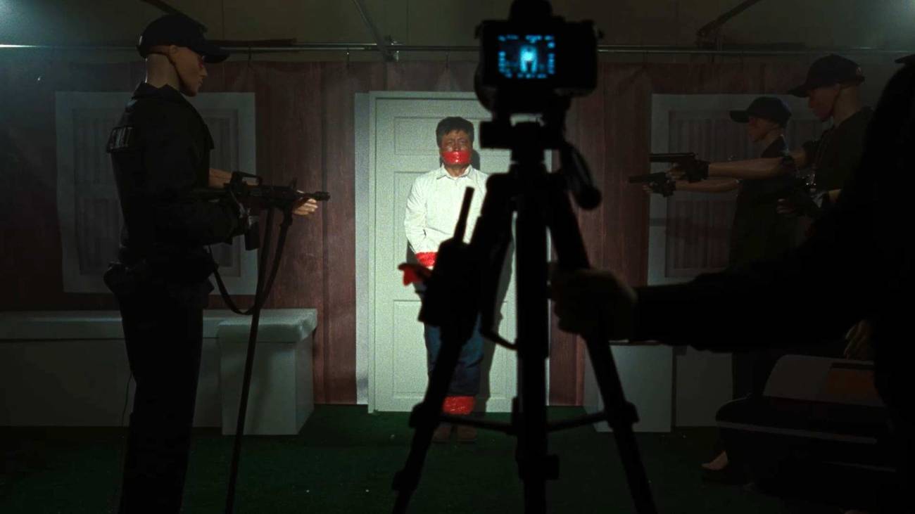 On a dimly lit basement stage with a video camera set up to record it, dummies are set up to represent a man outside of a door surrounded by cops with weapons, in a scene from Faces of Death (2026) recreating a scene from the 1978 film of the same name.