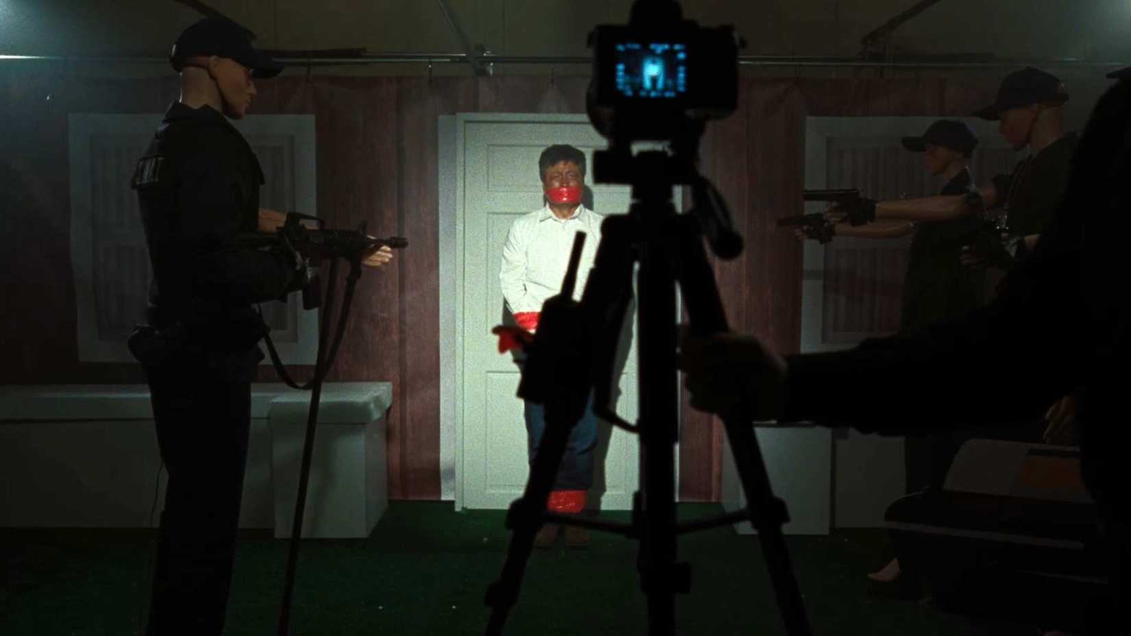 On a dimly lit basement stage with a video camera set up to record it, dummies are set up to represent a man outside of a door surrounded by cops with weapons, in a scene from Faces of Death (2026) recreating a scene from the 1978 film of the same name.