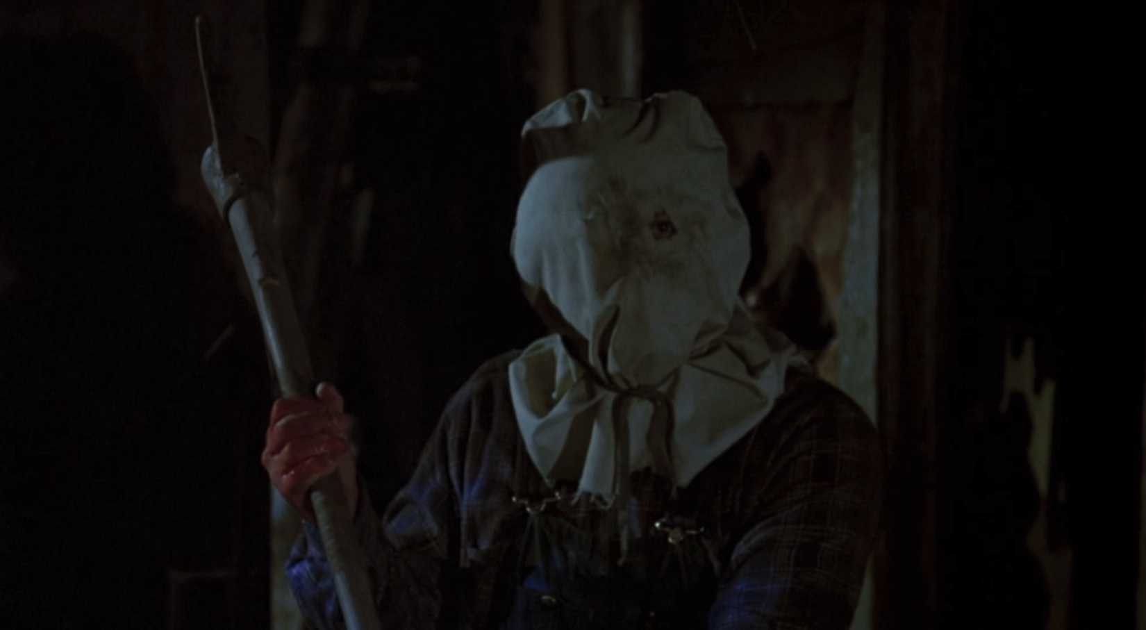 A man with a cloth sack over his head holds a pick axe threateningly