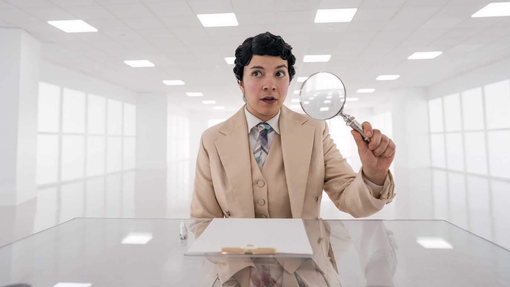 Muriel (Quelin Sepulveda), an angel wearing a tan suit and holding a magnifying glass sits in a stark white room in heaven in Good Opens.