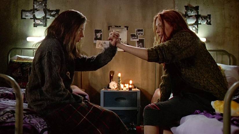 Two teen sisters, a brunette and a red-head, sitting on their respective beds, clasping hands