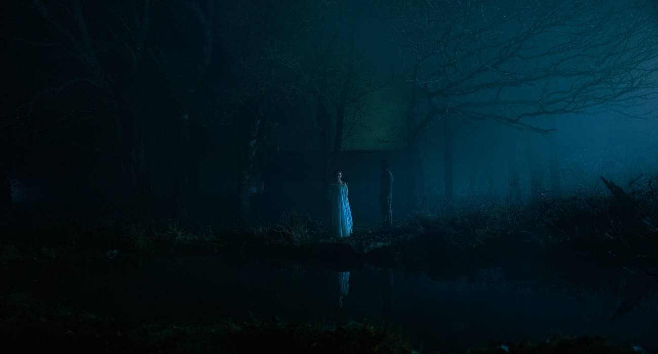 In a dark scene from The Green Knight, shot from a distance, a ghostly woman (Erin Kellyman) stands at the edge of a pond in the faint moonlight, with the knight Sir Gawain (Dev Patel) in shadow next to her.
