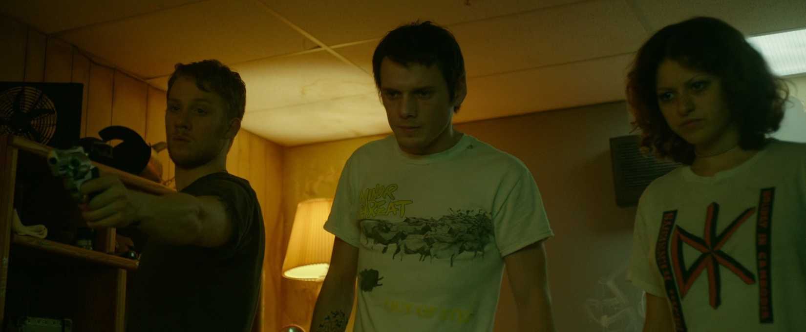 In a scene from the thriller Green Room, bandmates played by Joe Cole, Anton Yelchin, and Imogen Poots line up in the room where they've been trapped by neo-Nazi skinheads. The one played by Joe Cole, all the way to the left, brandishes a gun.