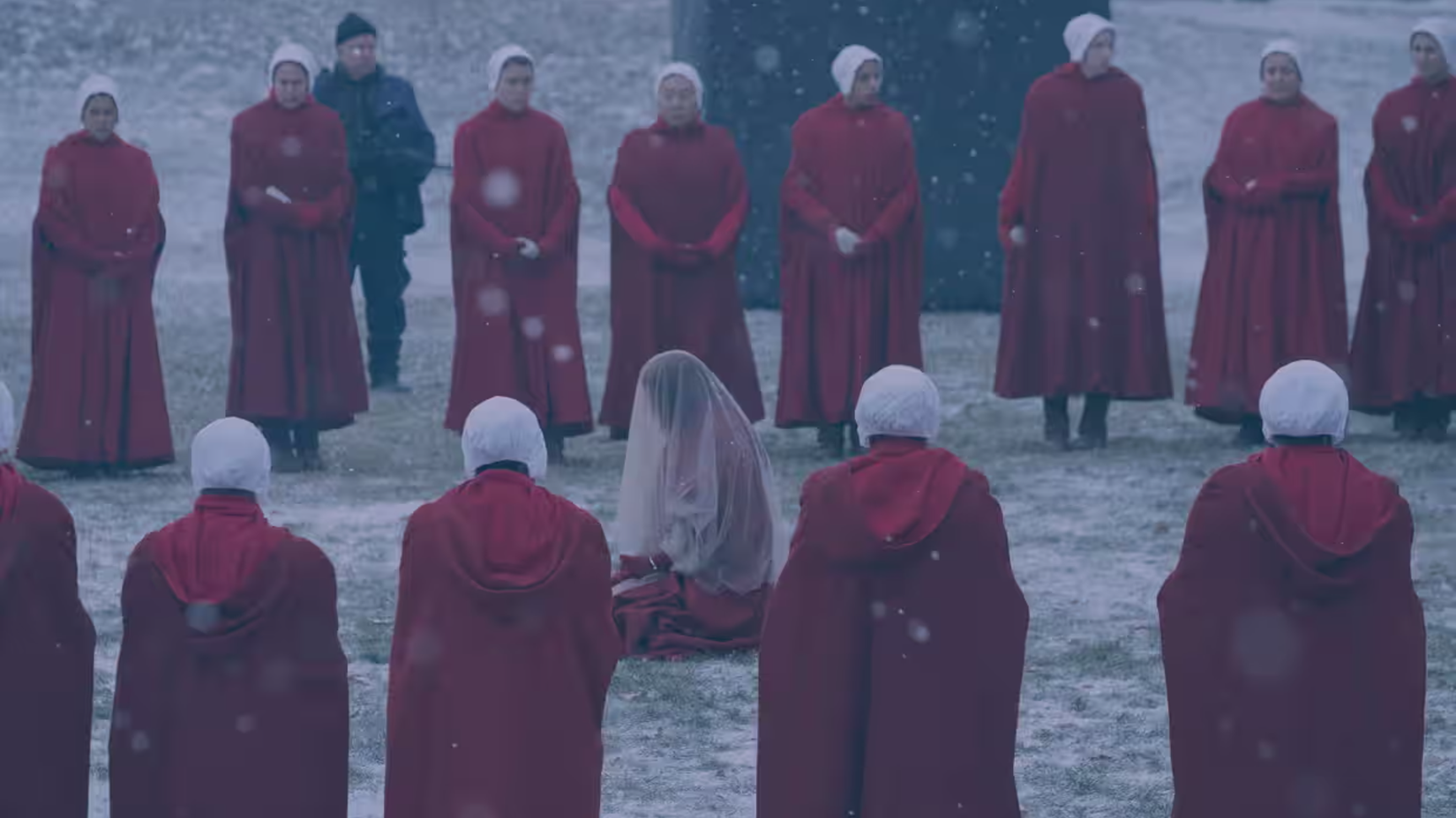 Women in red, hooded robes and gauzy white head-coverings stand in the snow in a circle around one of their number, who kneels in the snow wearing a white translucent veil in The Handmaid's Tale