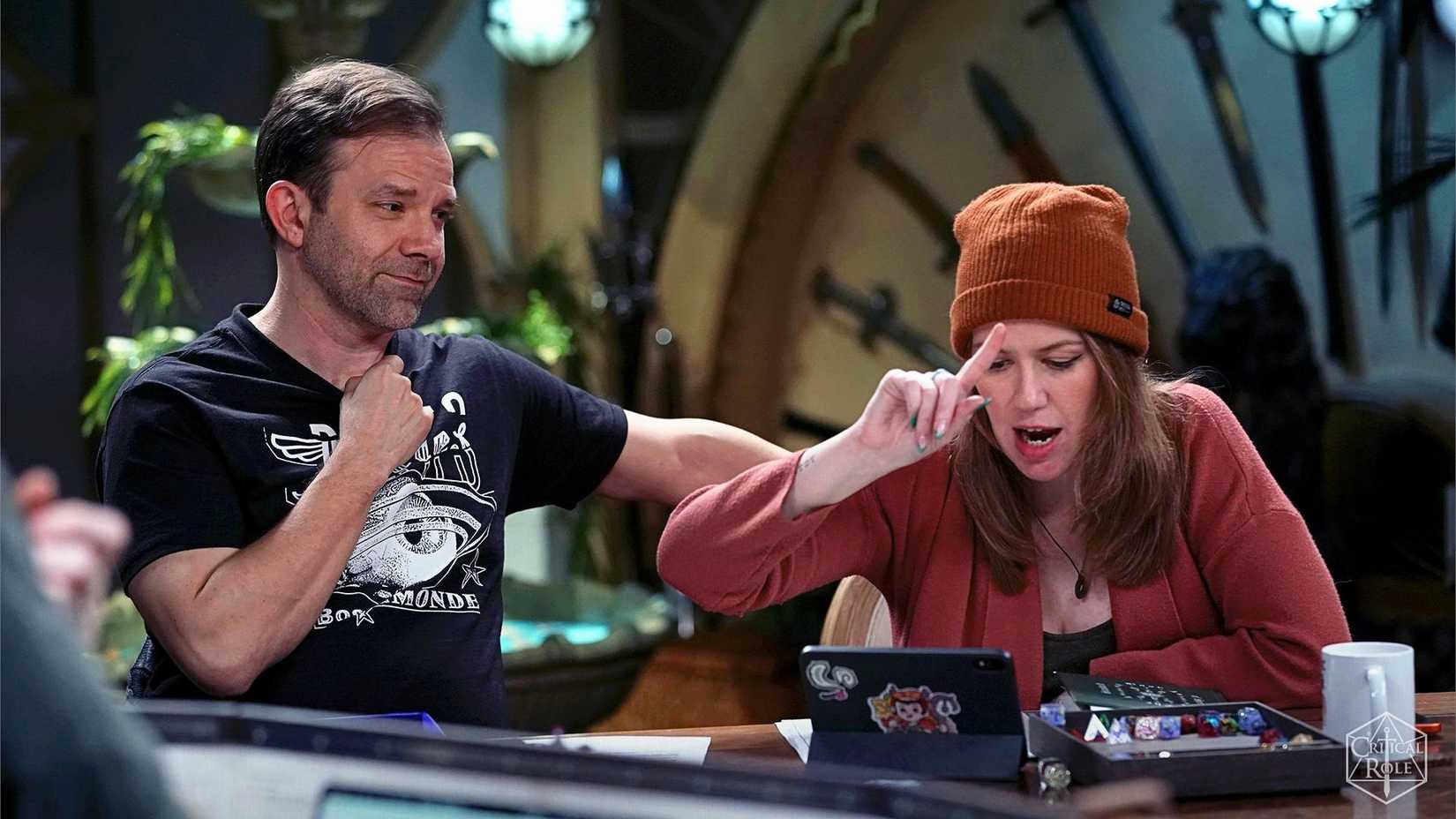 Critical Role image featuring Liam O'Brien and Marisha Ray looking at dice together. Marisha taps her head with a finger.